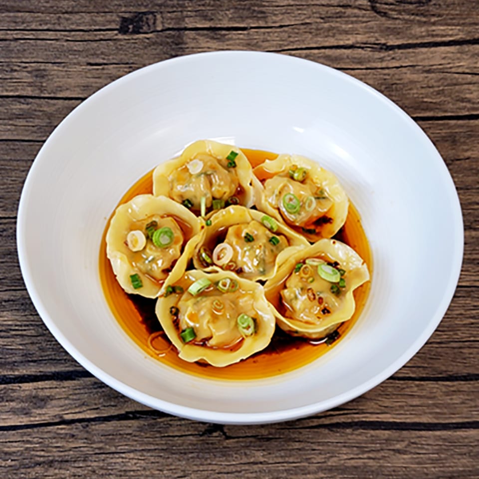 Six Brooklyn Dim Sum Co. dumplings in a white bowl, drizzled with sauce and garnished with scallions.