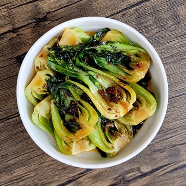 A white bowl filled with vibrant green bok choy, drizzled with a dark sauce, on a wooden table.