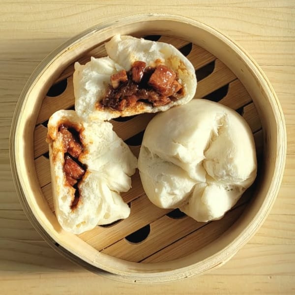Three char siu bao, two split open to show the savory pork filling, in a bamboo steamer.