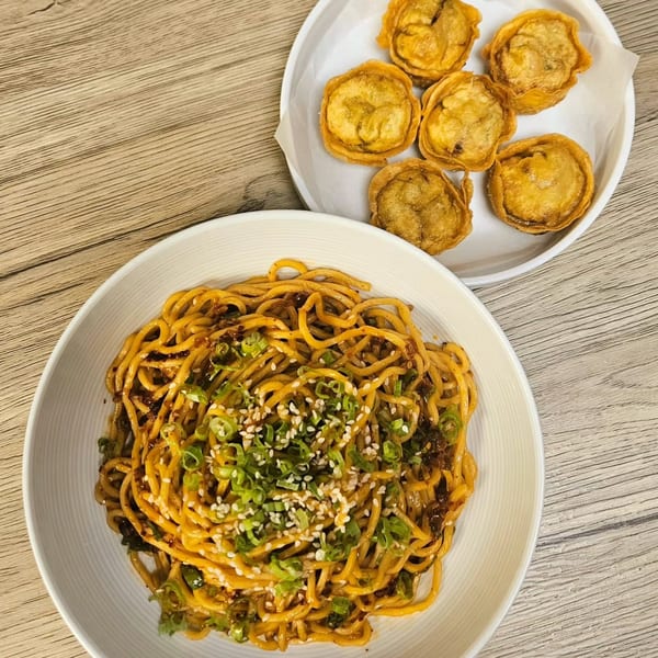 A bowl of sesame noodles with scallions and a plate of fried dumplings on a wooden table.