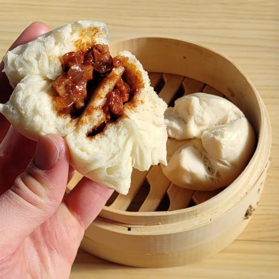 Hand holding a char siu bao, revealing its savory pork filling, with more buns in a bamboo steamer.