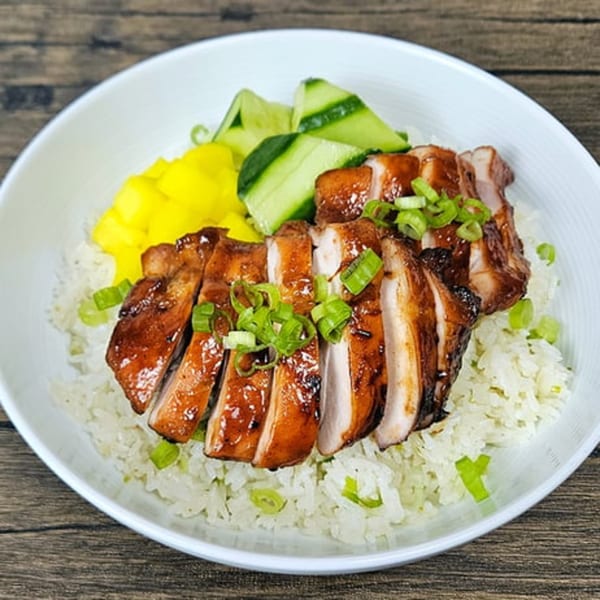 Teriyaki chicken, sliced and garnished with scallions, served over white rice with pickled cucumbers and mango.
