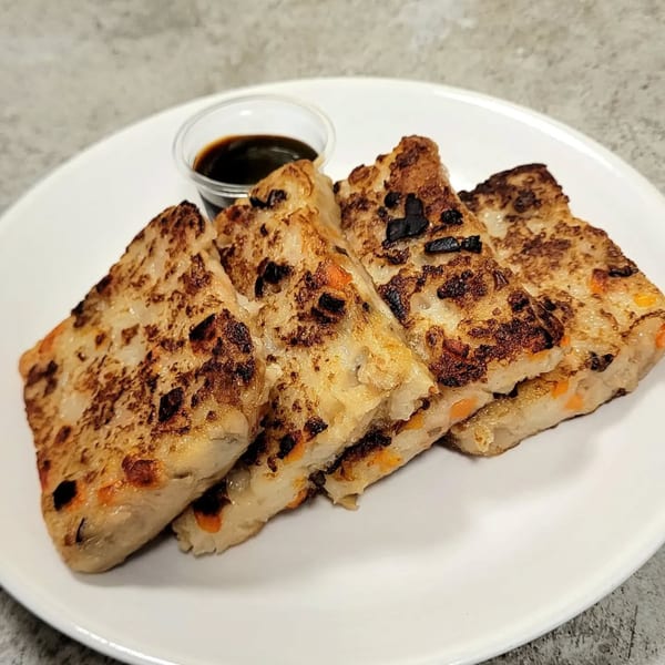 Four pan-fried turnip cakes with crispy edges and visible carrots, served with a side of soy sauce.