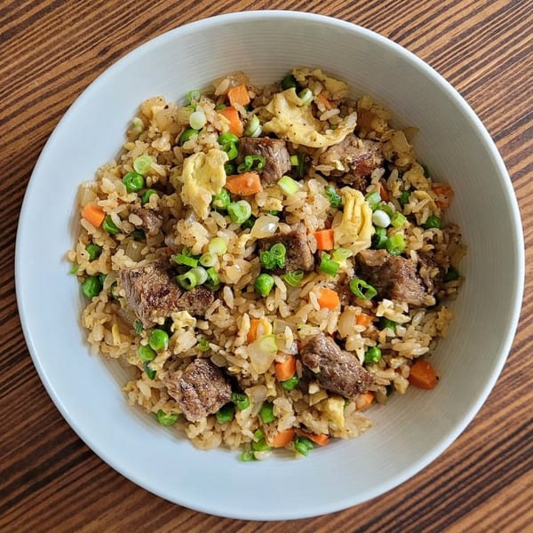 A white bowl filled with beef fried rice, peas, carrots, scrambled egg, and green onions.